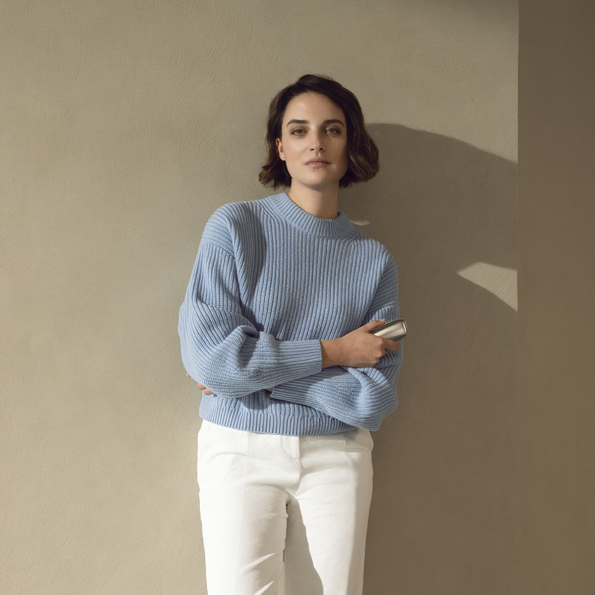 Woman with blue sweater standing against a wall, holding the Ploom X Advanced Silver device
