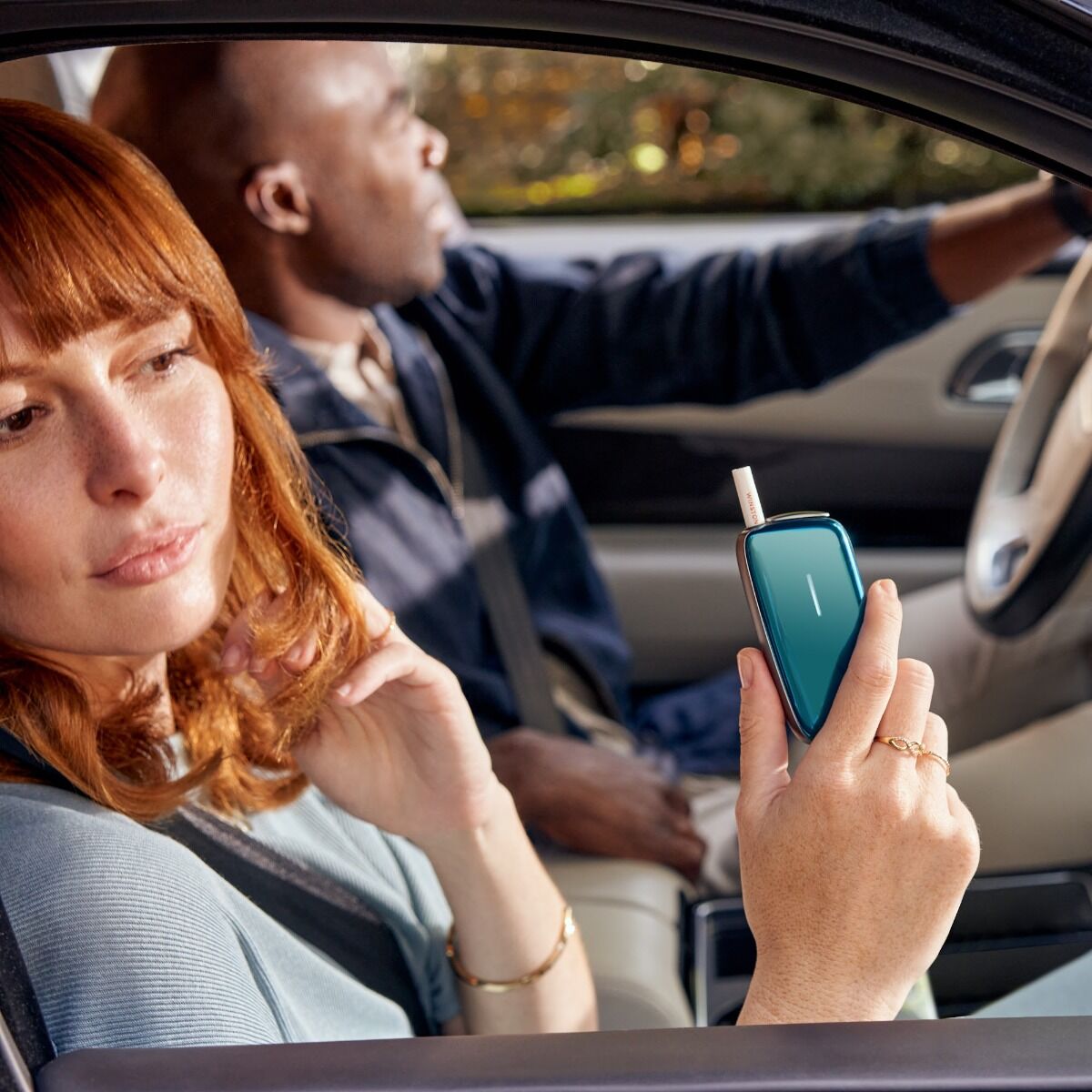 Woman in a car with a man, woman holding Ploom X Advanced with Ocean Blue front panel with tobacco stick, in hand