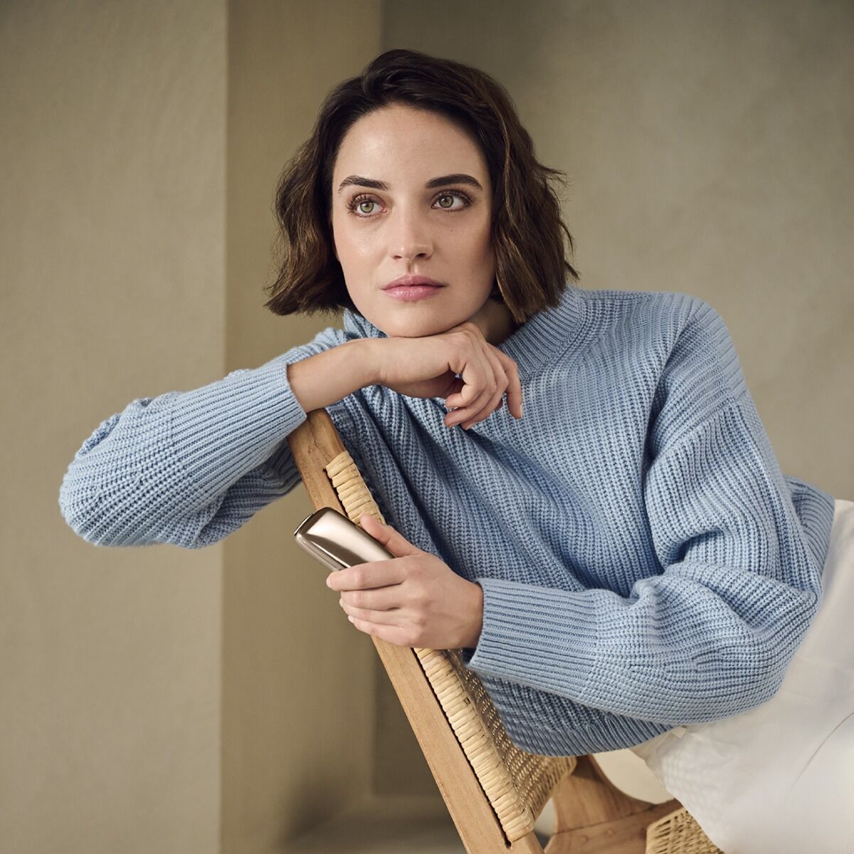 Mavi sviterli qadın kresloda oturub, əlində Ploom X Advanced Gold cihazı var