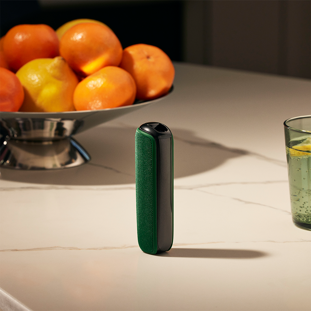 Meyvə səbətinin yaxınlığında masanın üzərində Amazon Green Arxa Örtüyü olan Ploom cihazı
