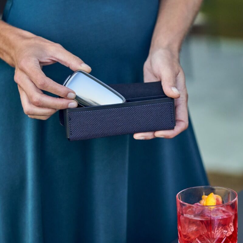 Woman in blue dress putting the Ploom device into a Ploom X Advanced Blue carry case, next to a drink
