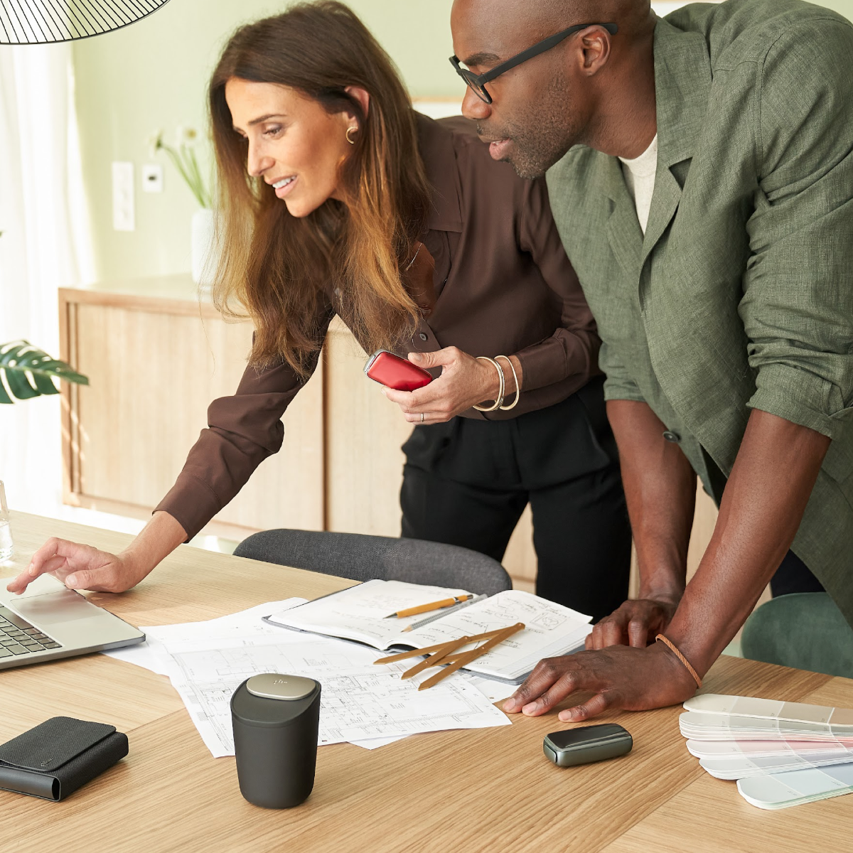 Man and woman working, leaning over a table, where the Ploom X Advanced large black stick ashtray, a black carry case and a Ploom device are displayed, along with a laptop, pens and paper and samples. The woman is holding a Ploom device with a Lava Red fr