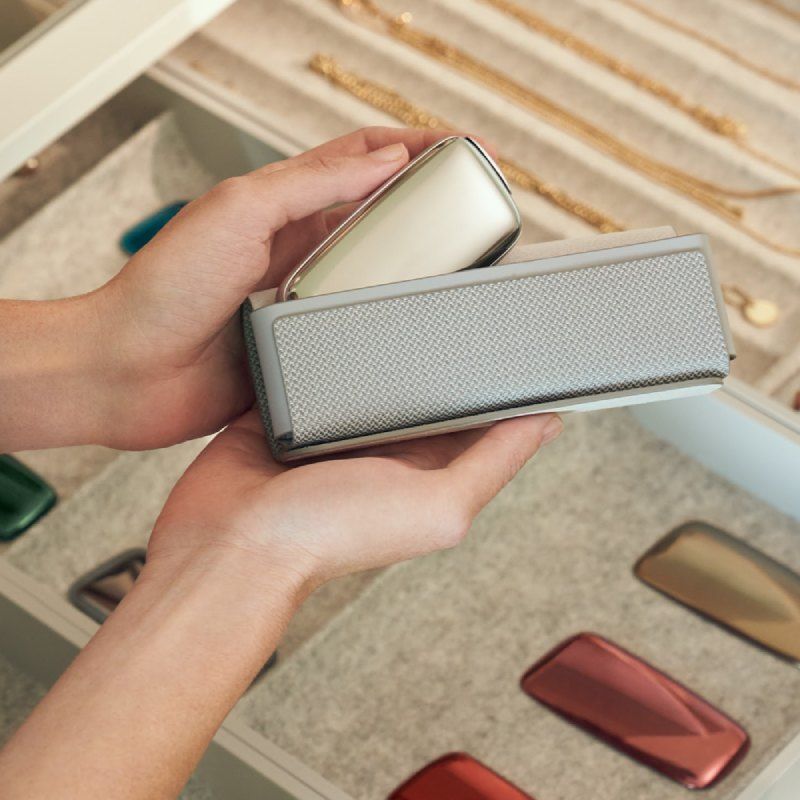 Ploom X Advanced Grey parça daşıma qutusu əlində tutulur — qızılı Ploom cihazı qutunun içinə yerləşdirilərkən, arxa planda zərgərlik əşyaları və Ploom ön panelləri olan siyirtmə fonunda göstərilir.